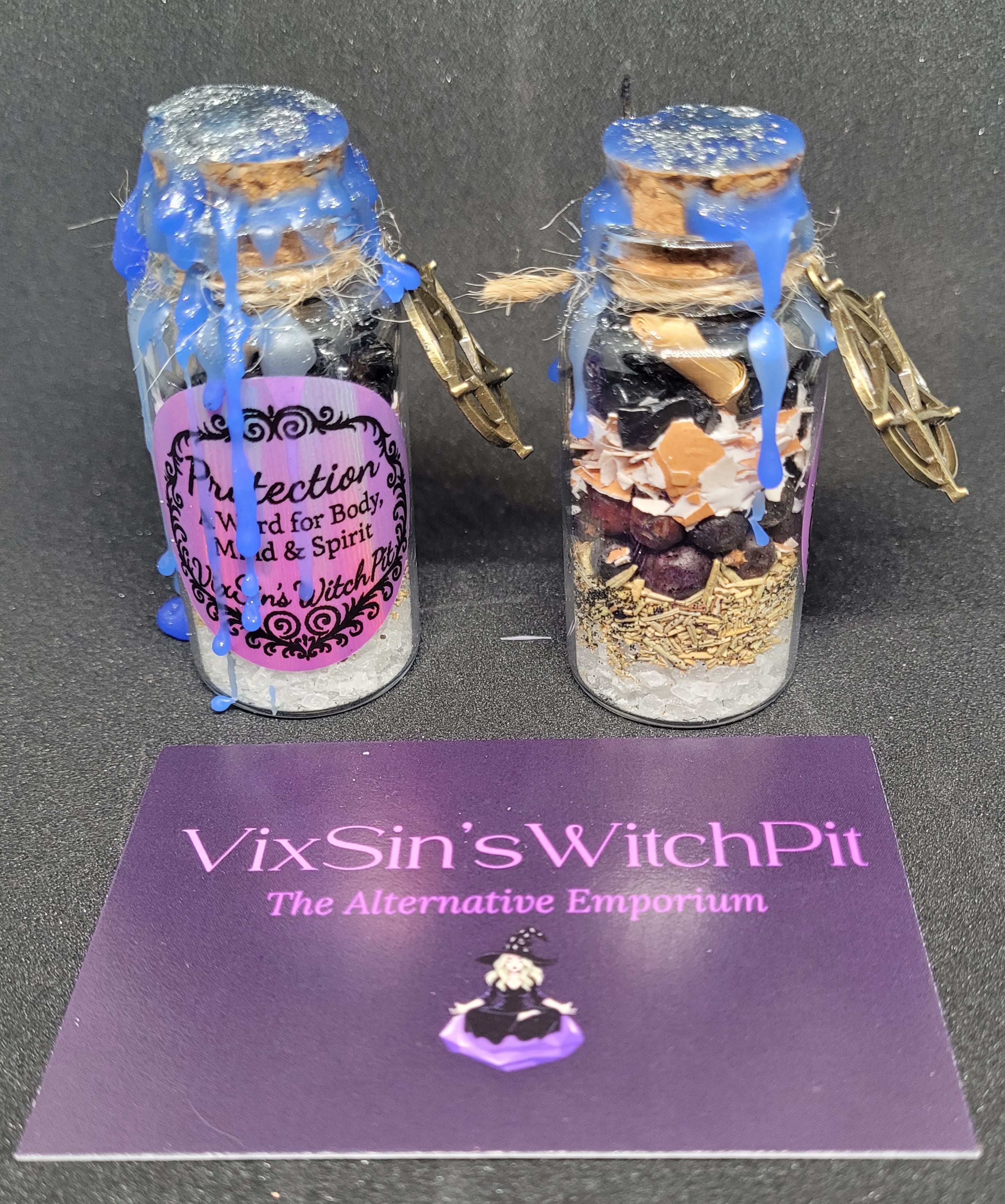 Two small glass Spell jars with decorative elements on a black background, featuring 'VixSin's WitchPit' branding. Protection.
