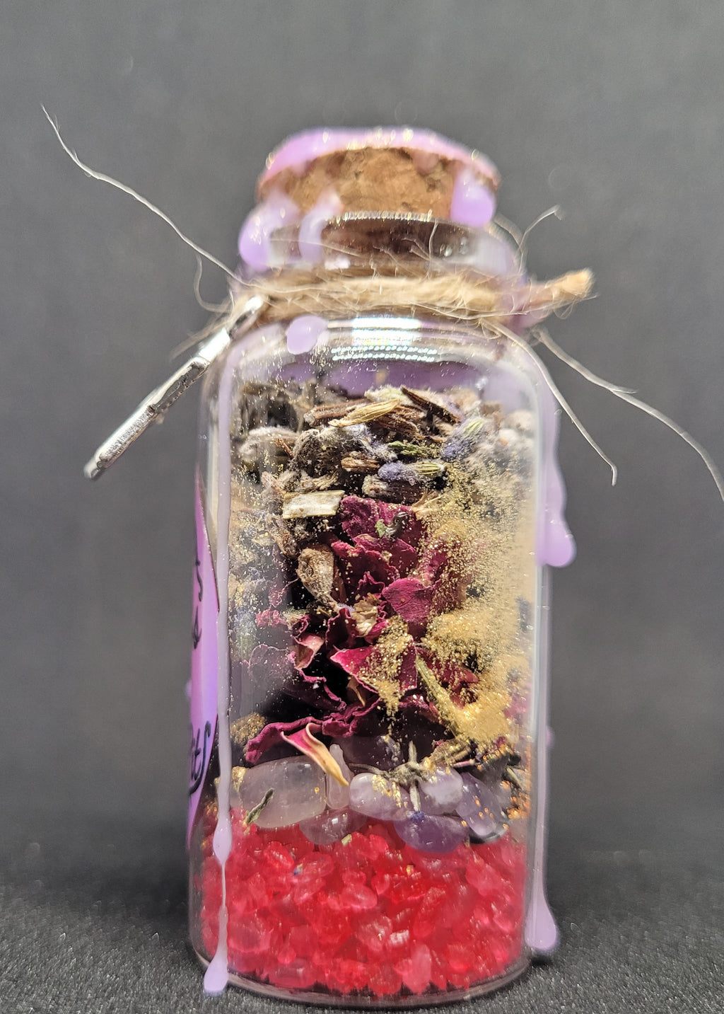 Small glass Spell-Jar with cork lid containing colourful dried flowers and leaves on a dark background
